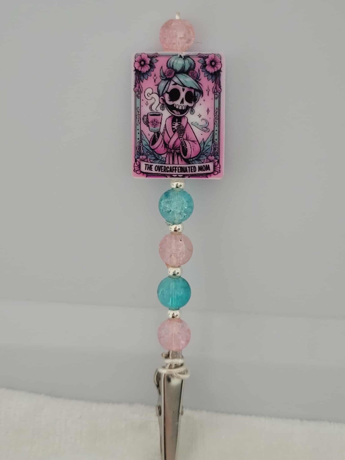 Decorative joint holder. Perfect for a mom. Roach clip with a silicone bead with an image of a pastel-punk skeleton mom in a pink robe with blue hair steaming cup of coffee, with words written that says "The Overcaffeinated mom", and paired with pretty pink and aqua crackled beads and small silver spacer beads.