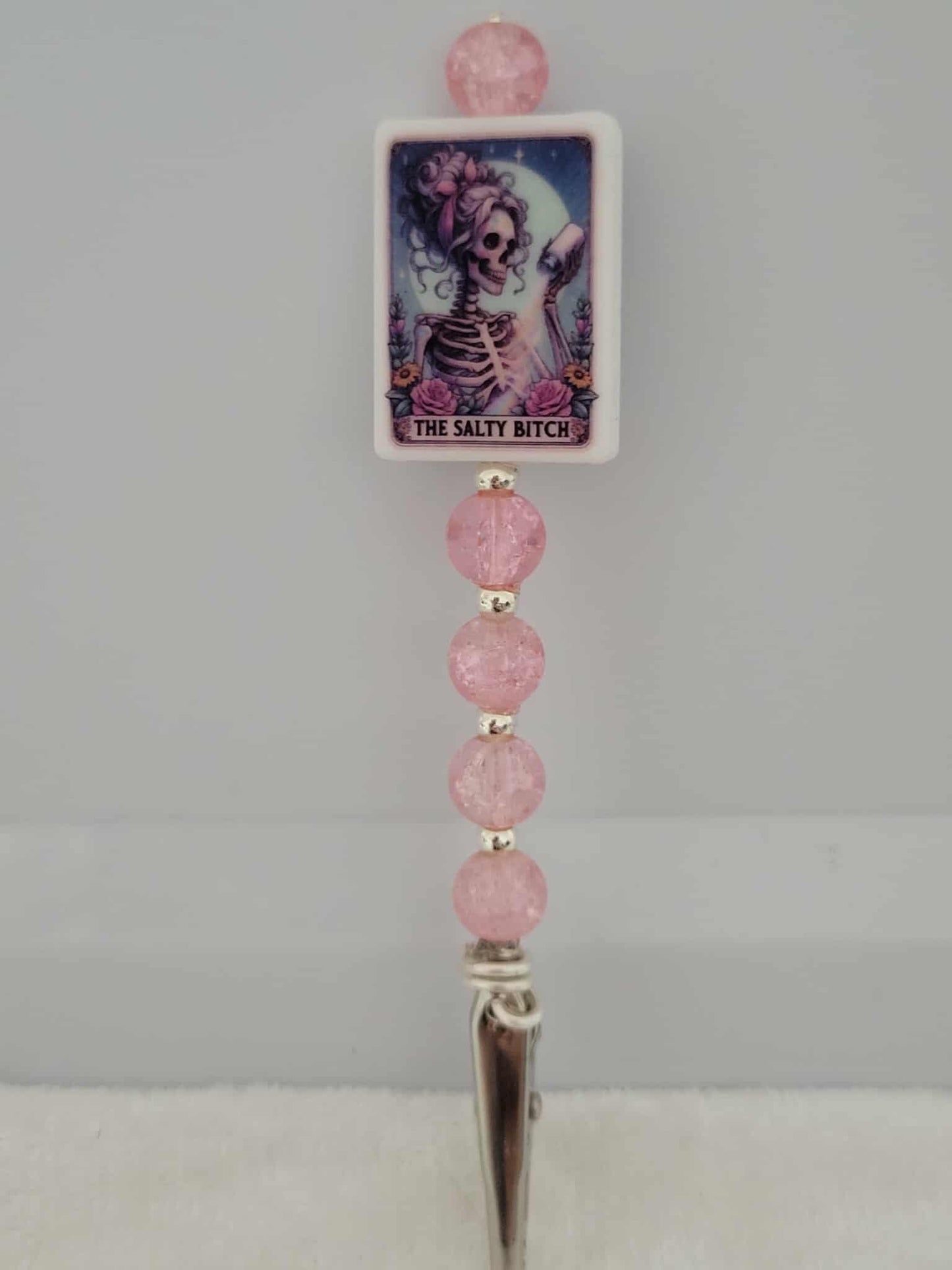 Decorative joint holder. Roach clip with a silicone bead that has an image of a skeleton babe with messy curly hair, tossing salt under the moonlight, that reads "The Salty Bitch", along with pink crackle beads and small silver beaded spacers.
