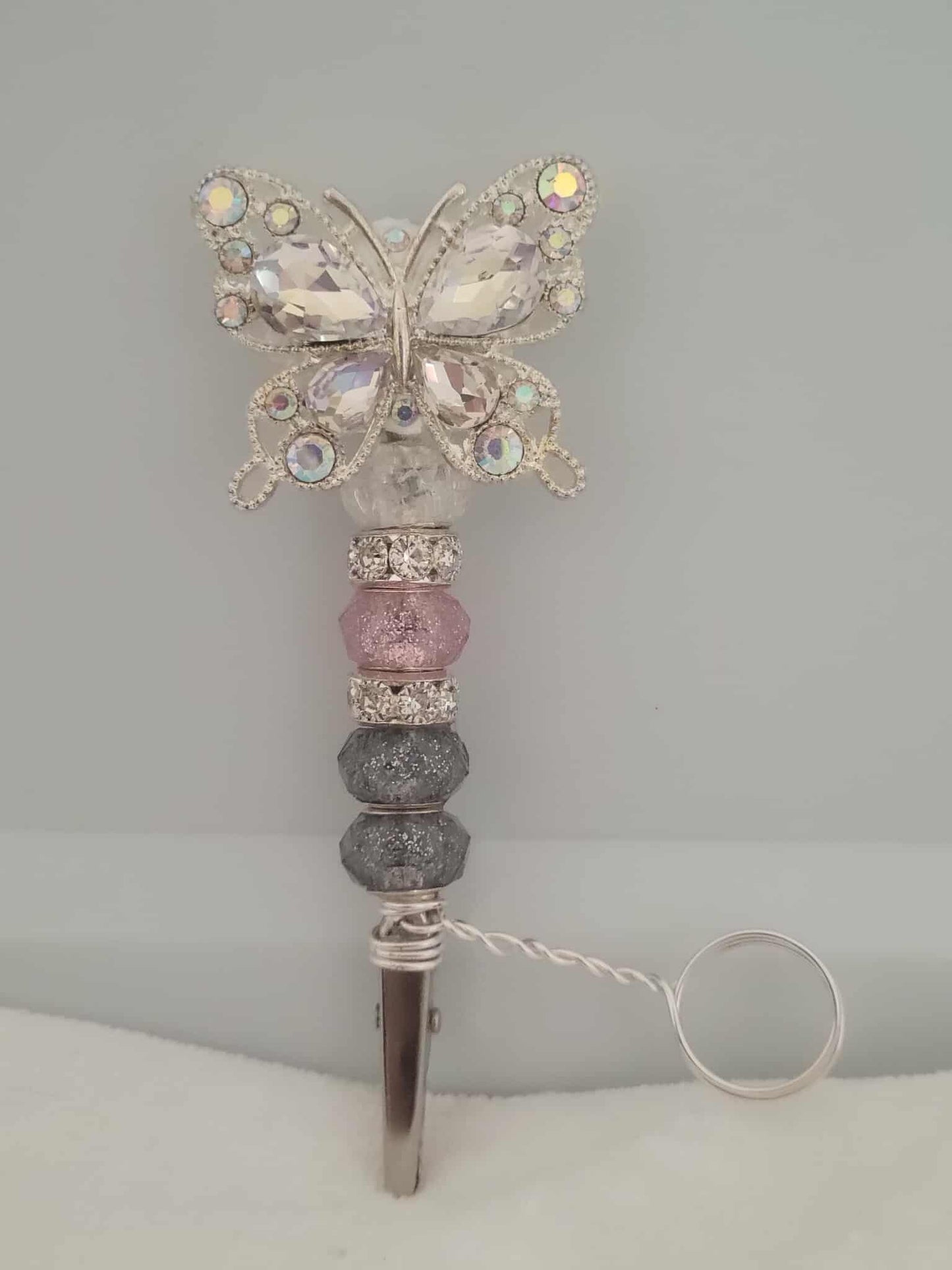 Decorative joint holder. Sparkling crystal butterfly roach clip with pink glitter-dipped beads, and a sleek silver beaded grip. There is a metal ring extending from right above the roach clip and below the start of the beads. This allows users to wear the ring on their finger that holds the roach clip allowing the user to hold other objects like your phone or a video game controller. The ring is not adjustable and was made to be worn as a midi-ring, about a midi ring size 3 (14mm).