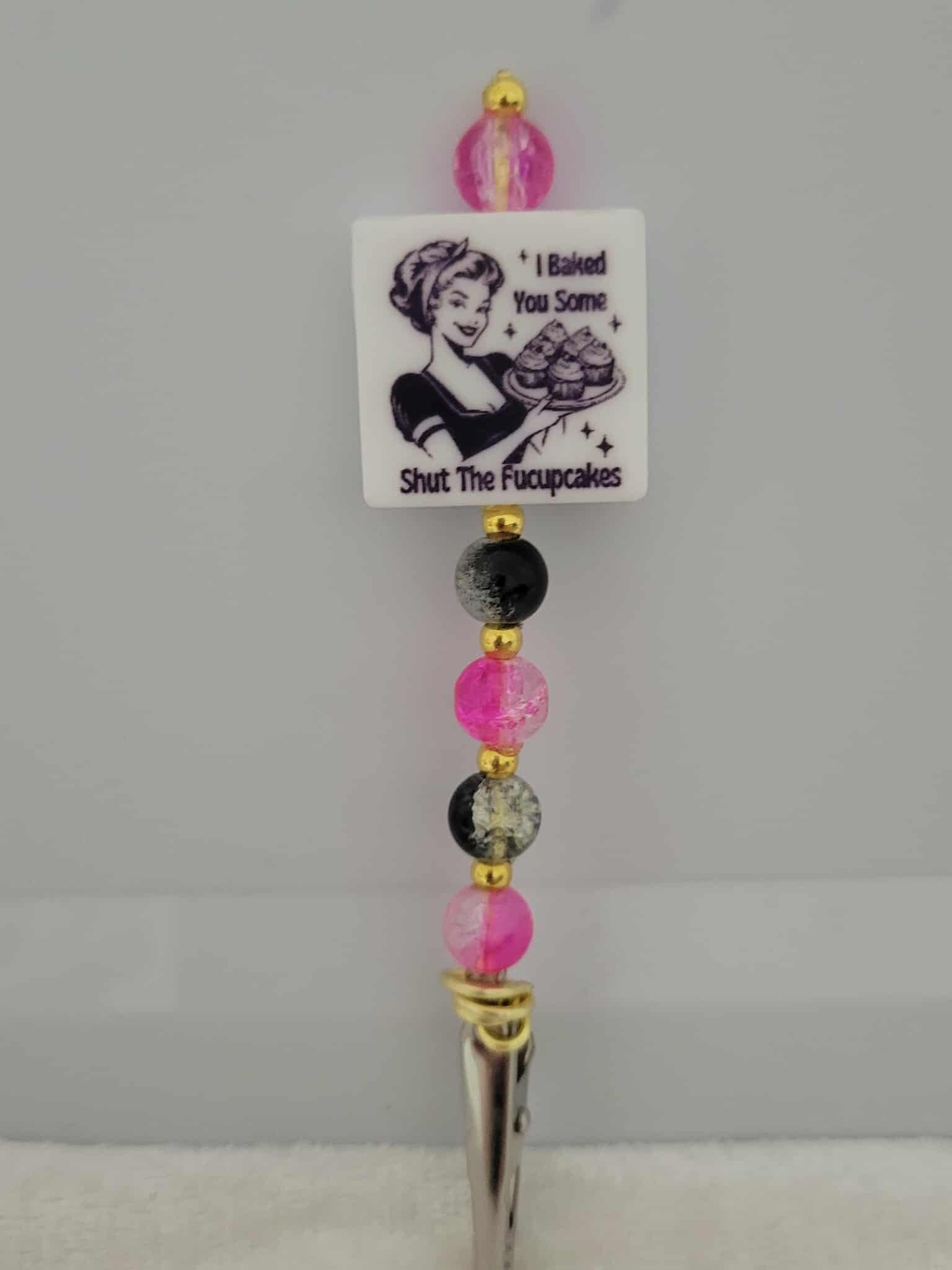 Decorative joint holder. A roach clip with a silicone bead with an image of a retro housewife holding cupcakes and the message, “I baked you some shut the f*ckupcakes,”, along with pink and black crackle beads and small gold spacers.