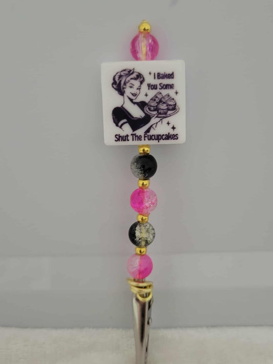 Decorative joint holder. A roach clip with a silicone bead with an image of a retro housewife holding cupcakes and the message, “I baked you some shut the f*ckupcakes,”, along with pink and black crackle beads and small gold spacers.