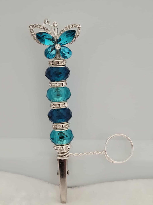 Handcrafted silver and blue butterfly roach clip, with sparkling rhinestone spacers. There is a metal ring extending from right above the roach clip and below the start of the beads. This allows users to wear the ring on their finger that holds the roach clip allowing the user to hold other objects like your phone or a video game controller.  The ring is not adjustable and was made to be worn as a midi-ring, about a midi ring size 3 (14mm)