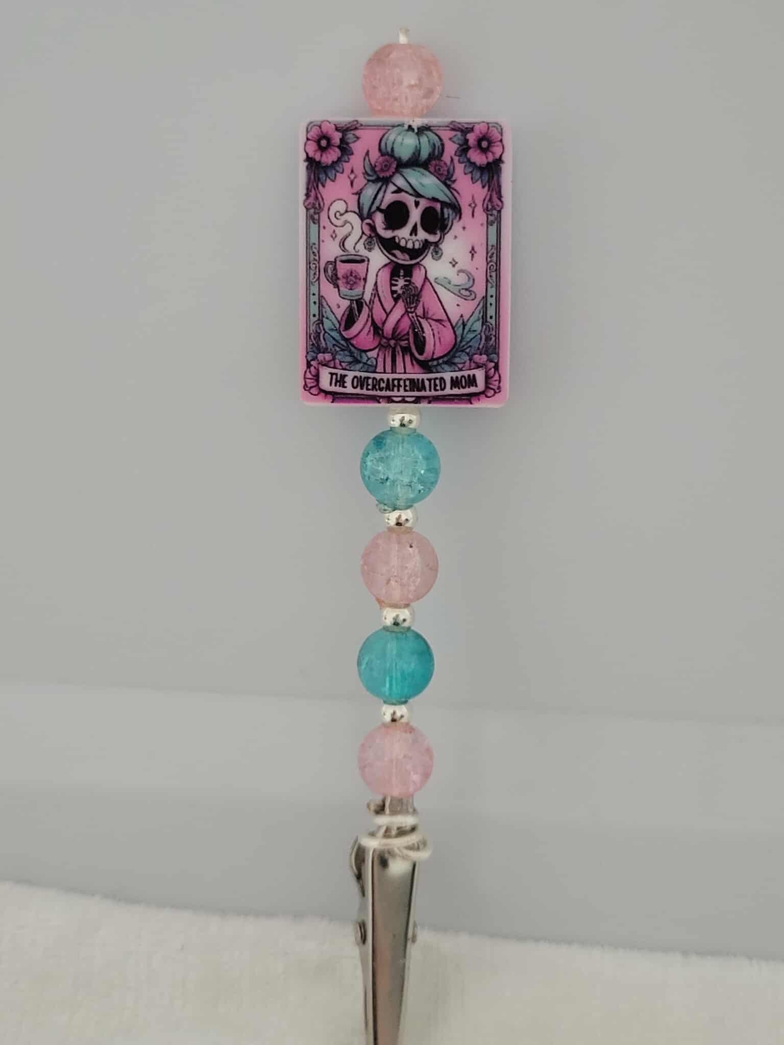 Decorative joint holder. Perfect for a mom. Roach clip with a silicone bead with an image of a pastel-punk skeleton mom in a pink robe with blue hair steaming cup of coffee, with words written that says "The Overcaffeinated mom", and paired with pretty pink and aqua crackled beads and small silver spacer beads.