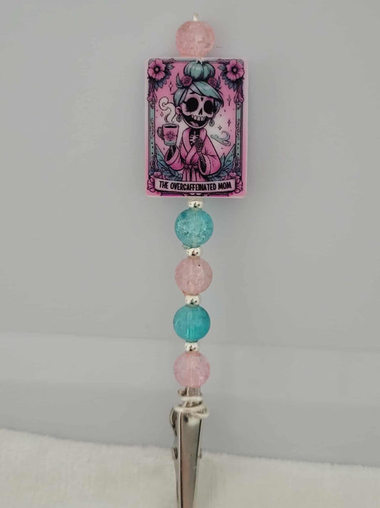 Decorative joint holder. Perfect for a mom. Roach clip with a silicone bead with an image of a pastel-punk skeleton mom in a pink robe with blue hair steaming cup of coffee, with words written that says "The Overcaffeinated mom", and paired with pretty pink and aqua crackled beads and small silver spacer beads.