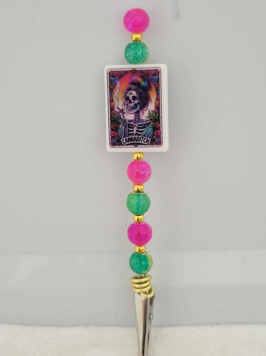Decorative joint holder. Roach clip with a silicone bead with an image of a rainbow-lit skeleton girl smoking surrounded by cannabis leaves and flowers, with the words written "CANNABITCH". Along with hot pink and green crackle beads with gold accents.