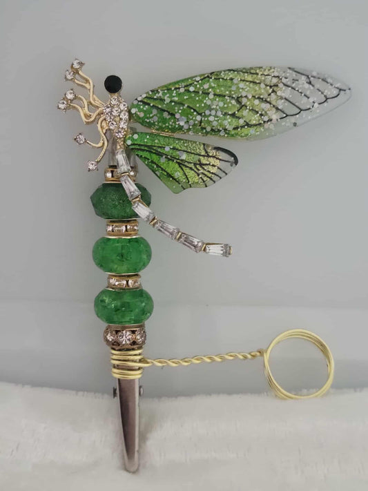 Decorative joint holder. Gold dragonfly roach clip with green wings, shimmering emerald beads, dazzling gold accents, and a convenient side ring allows you to keep playing your video games or scrolling on your phone. The ring is not adjustable and was made to be worn as a midi-ring, about a midi ring size 3 (14mm).