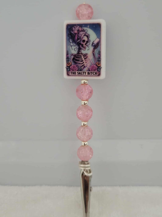 Decorative joint holder. Roach clip with a silicone bead that has an image of a   skeleton babe with messy curly hair, tossing salt under the moonlight, that reads "The Salty Bitch", along with pink crackle beads and small silver beaded spacers.