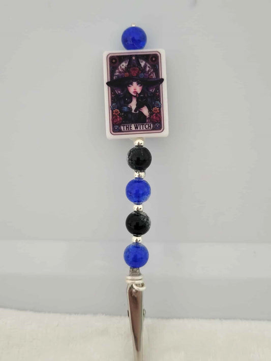 Decorative joint holder. Roach clip with a silicone bead with an image of a young gothic girl witch surrounded by red flowers, with the words written that says “The Witch "along with black and deep sapphire crackle beads and small silver spacers.